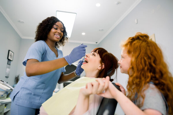 Side bottom view of young Caucasian woman having teeth checkup and treatment. Her little daughter holds hand of mom, while female black dentist with tools provides teeth cure Side bottom view of young Caucasian woman having teeth checkup and treatment. Her little daughter holds hand of mom, while female black dentist with tools provides teeth cure.
