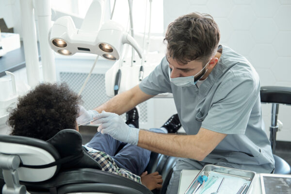 Dentist bending over little boy while drilling his tooth Dentist in uniform, glovea and protective mask bending over little boy while drilling his tooth after examination of oral cavity